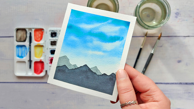 kleine Aquarell-Landschaft mit blauem Wolkenhimmel und Bergen