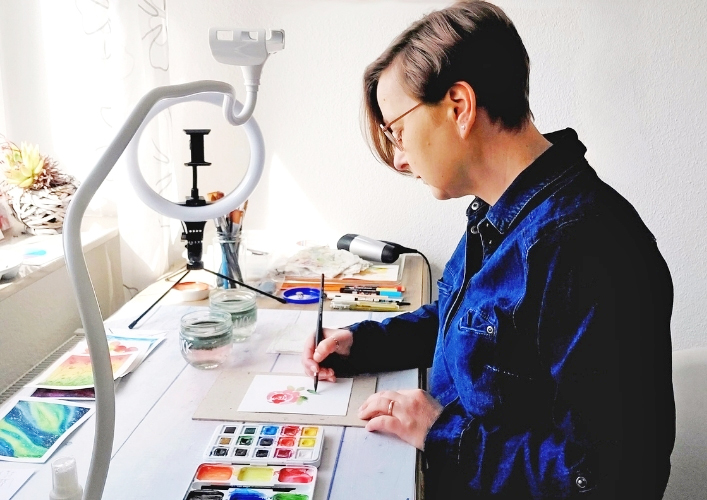 Frau in blauem Overall sitzt an Malplatz und malt