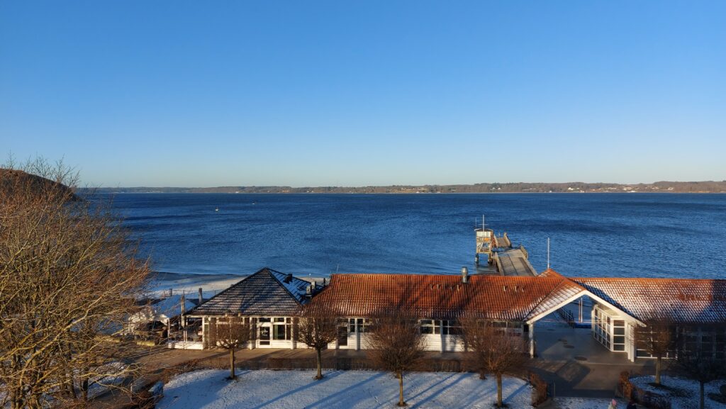 Blick auf windgepeitschtes Wasser im Vordergrund flache Gebäude mit roten Dächern