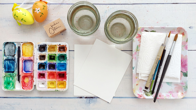 Aquarellfarbkasten, Aquarellpapier, Pinsel, Wasserbecher auf Malplatz plus Osterdeko