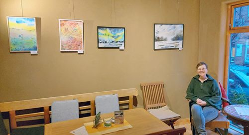 Frau sitzt auf Stuhl, an der Wand vier Aquarell-Bilder mit Galerie-Aufhängung