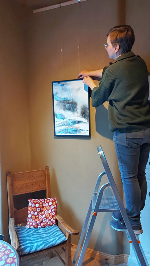 Frau steht auf Leiter und hängt Aquarellbild auf