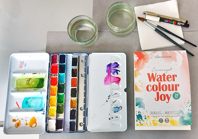Malplatz mit Aquarellfarbkasten, Wasserbechern, Pinseln und Papierblock auf Glastisch